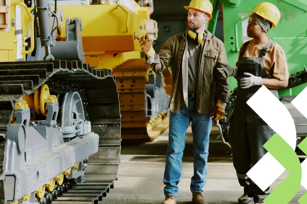 Dois técnicos de manutenção, um homem e uma mulher, equipados com capacetes, inspecionando a esteira de um trator amarelo em uma oficina.