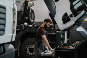 Técnico de manutenção seleciona ferramentas em oficina de frota pesada, com dois caminhões em revisão preventiva e cabine basculada ao fundo
