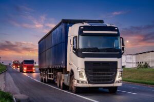 Comboio de caminhões de frota rodoviária trafegando em rodovia ao entardecer, com caminhão branco em primeiro plano e caminhões vermelhos ao fundo.